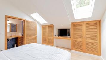 Ein Schlafzimmer mit einem Bett, Dachfenstern, einem Fernseher, Holzschränken und einem Badezimmer mit Waschbecken und Spiegel.