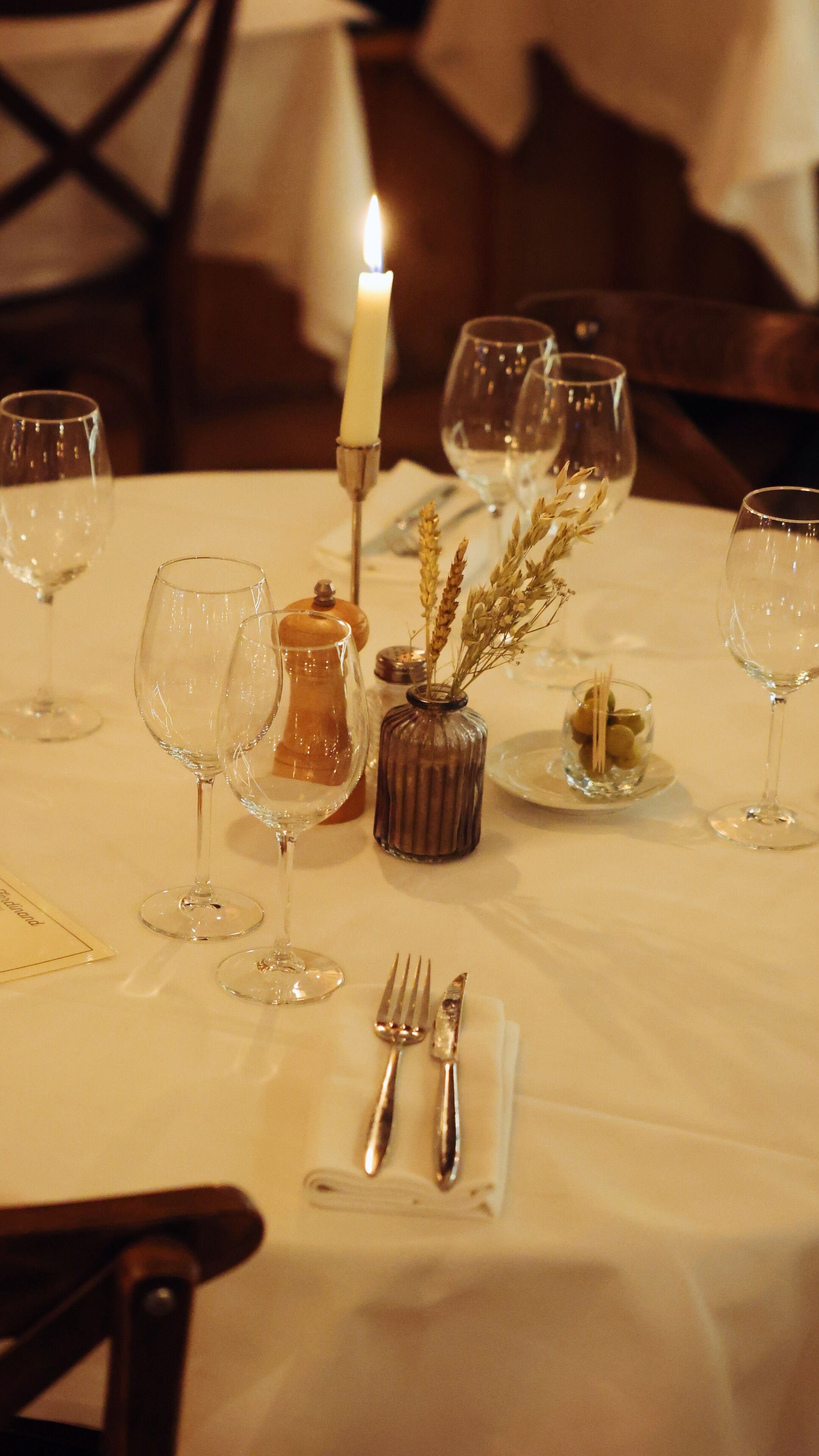 Una mesa dispuesta para una comida, con copas de vino, una vela y un pequeño jarrón con flores secas.