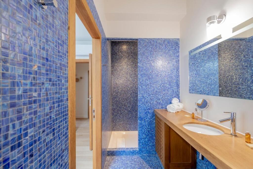 A modern bathroom with blue mosaic tiles, a wooden vanity, a large mirror, and a shower area.