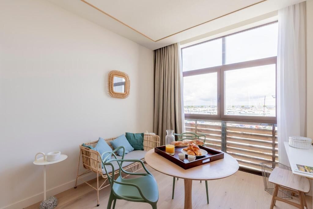 A modern hotel room with a large window, a round dining table with breakfast, a wicker sofa, and a small side table.