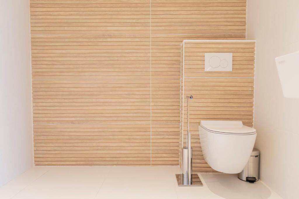 Un baño moderno con pared de paneles de madera, inodoro blanco y lavamanos de acero inoxidable.