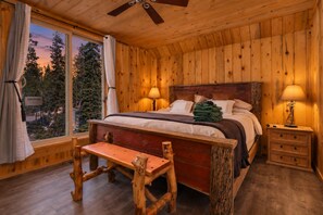 A wooden cabin bedroom with a bed, bedside lamps, a bench, and a window with a view of trees.