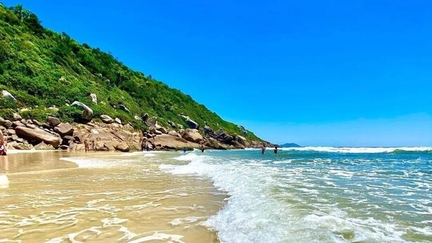 A beach with clear waters, a rocky shoreline, and a green hillside.