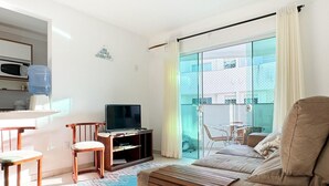 A modern living room with a sofa, a TV, a dining table with chairs, and a large window with curtains.