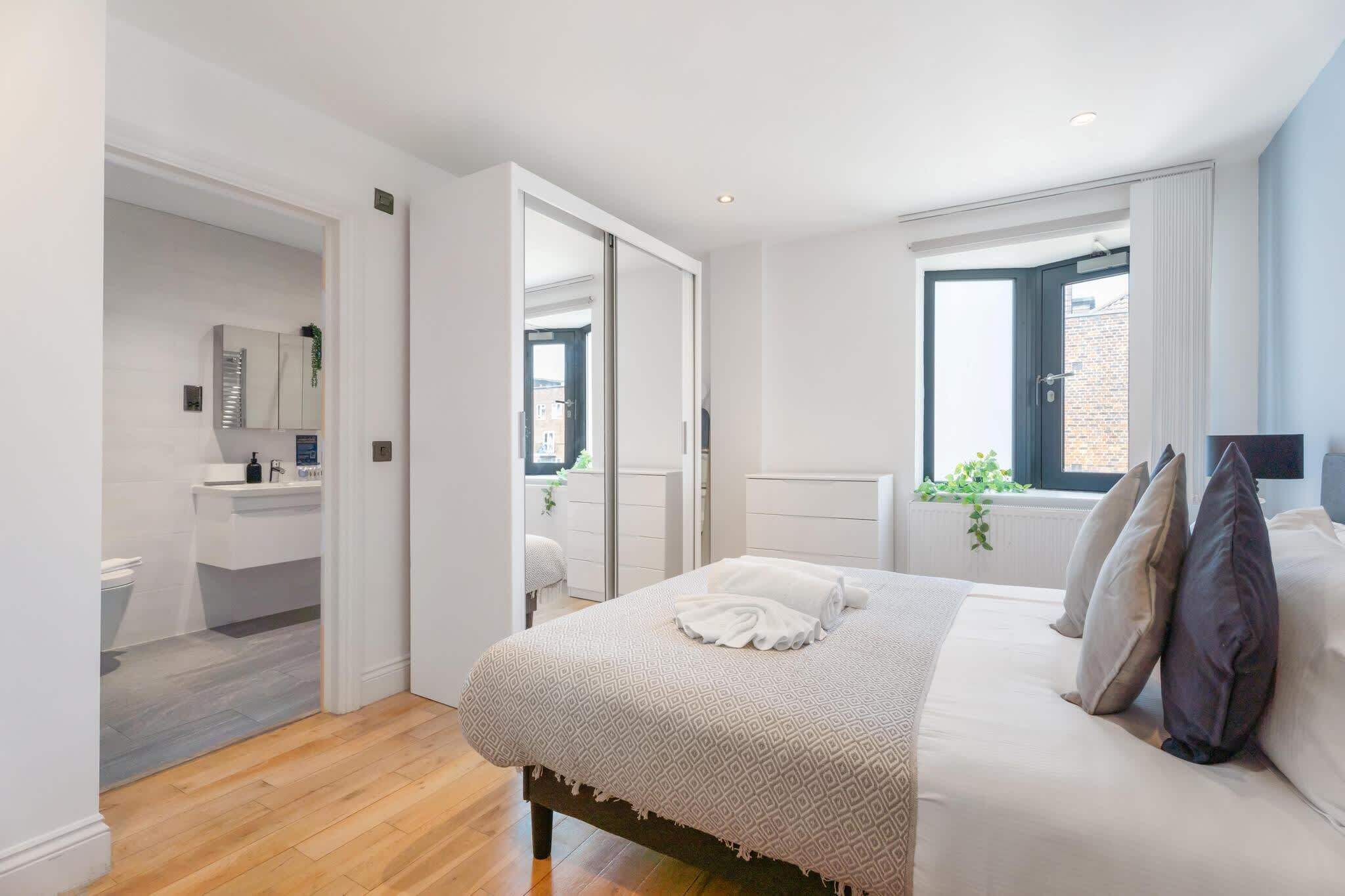 A bedroom with a bed, a window, a plant, and a bathroom with a sink and mirror.