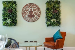 A room with a round table, a single orange chair with a blue pillow, and two vertical green wall decorations.