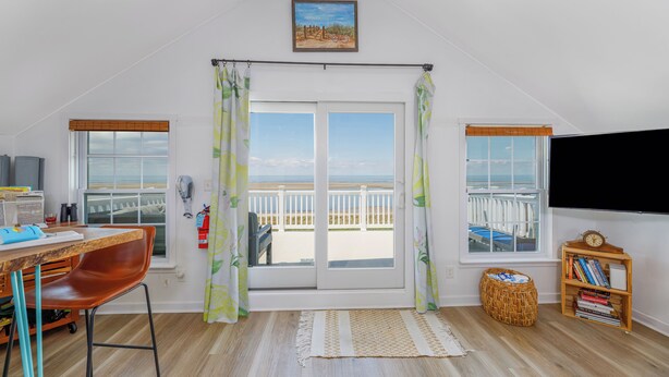 Ein Zimmer mit Schreibtisch aus Holz, einem Sessel, einem Fernseher, einem Bücherregal und einem Balkon mit Blick auf den Strand.