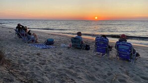 Ein Sonnenuntergangsblick über einen Strand, mit Menschen, die auf Stühlen zum Meer sitzen.