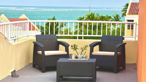 Ein Balkon mit zwei Korbstühlen und einem kleinen Tisch, mit Blick auf einen Strand mit Palmen und eine Meerblick.