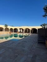 Zona de piscina con cielo azul despejado, suelo de baldosas y un edificio con arcos al fondo.