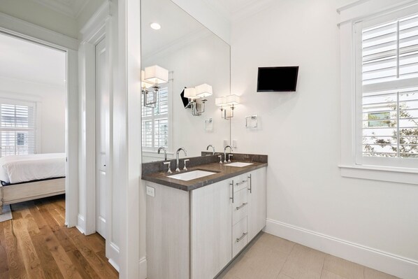 A modern bathroom with two sinks, a large mirror, and a flat-screen TV mounted on the wall.