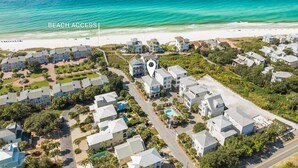 An aerial view of a coastal residential area with beachfront access, featuring a mix of single and multi-story houses, a central road, and a clear view of the ocean.