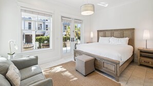 A bedroom with a large bed, a sofa, a small ottoman, a nightstand, and a lamp. There is a window with a view of the outside.
