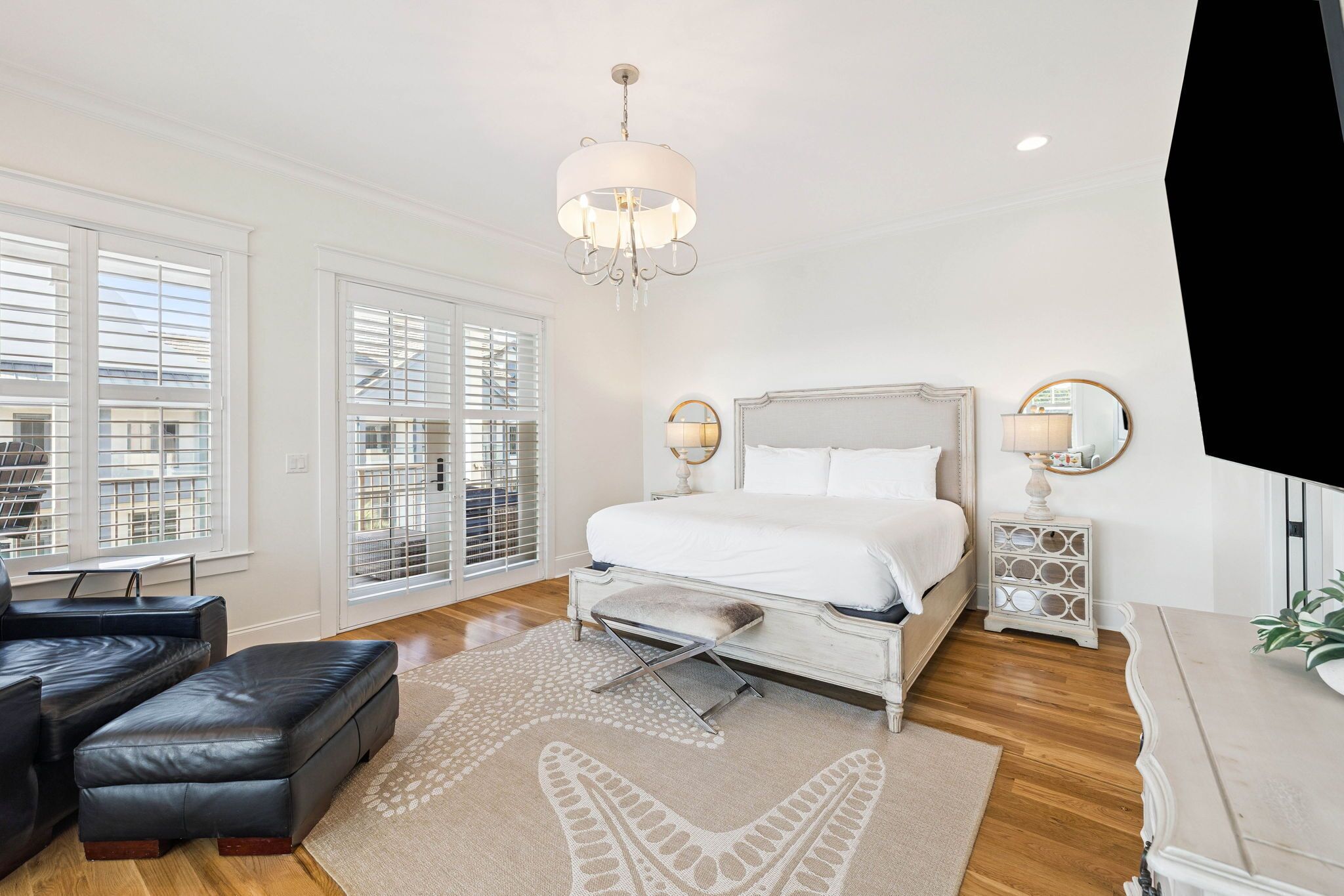 A bedroom with a large bed, a chandelier, a bedside table with a mirror, and a view of the outside through the window.