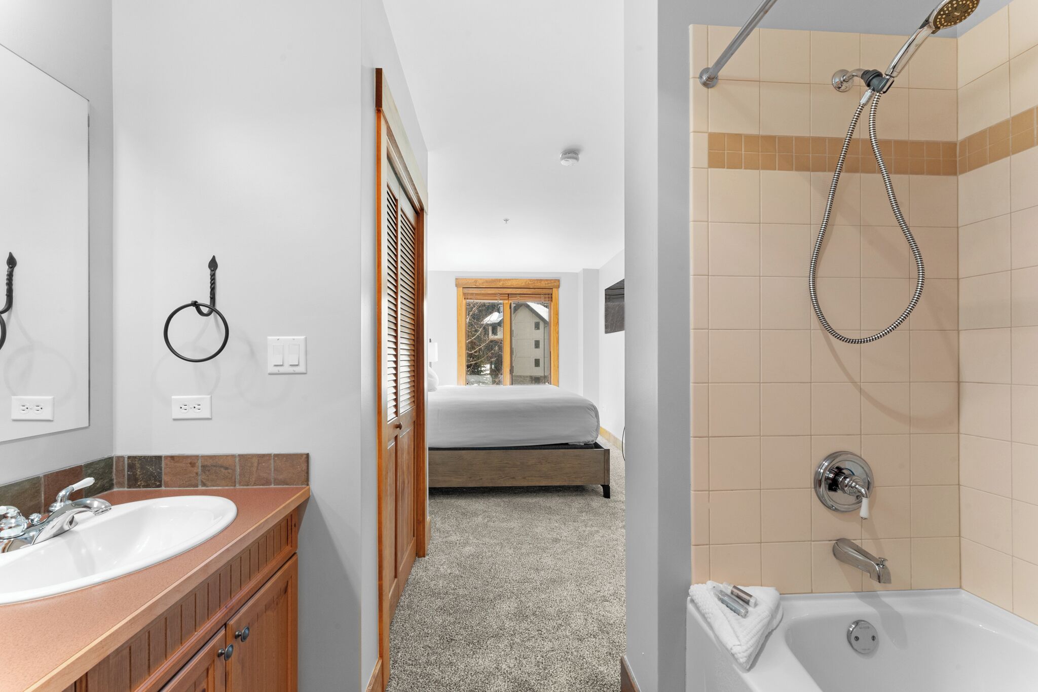 A bathroom with a sink, shower, and a bed visible through an open door.