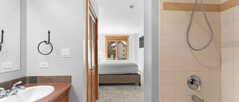 A bathroom with a sink, shower, and a bed visible through an open door.