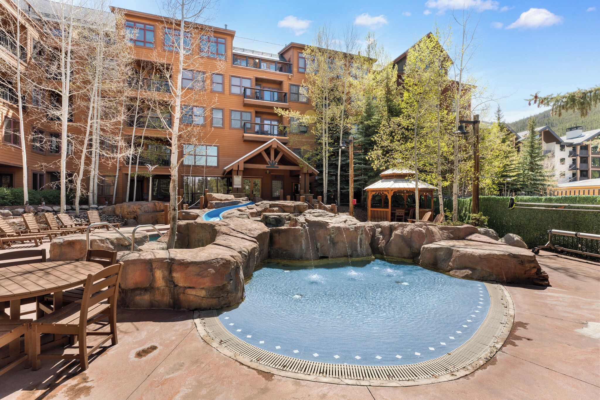 A pool area with wooden lounge chairs, a hot tub, and a building with multiple balconies.