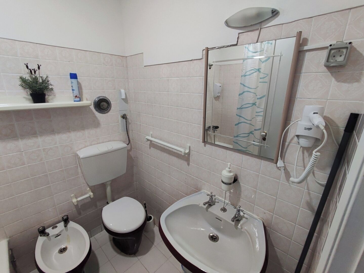 A small bathroom with a toilet, bidet, and sink, a mirror, and a shelf with toiletries.