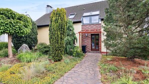 Een huis met een stenen pad dat leidt naar een houten deur, omringd door verschillende planten en bomen.