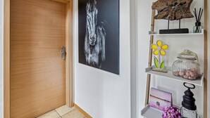 Een gang met een houten deur, een zwart-wit paardenportret en een plank met decoratieve objecten, waaronder een bloem, een pot en een kleine plant.