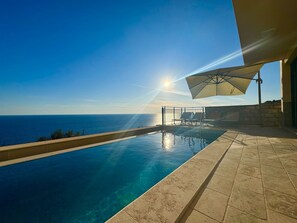 Una piscina infinita con vista clara al océano, una sombrilla y un muro de piedra.