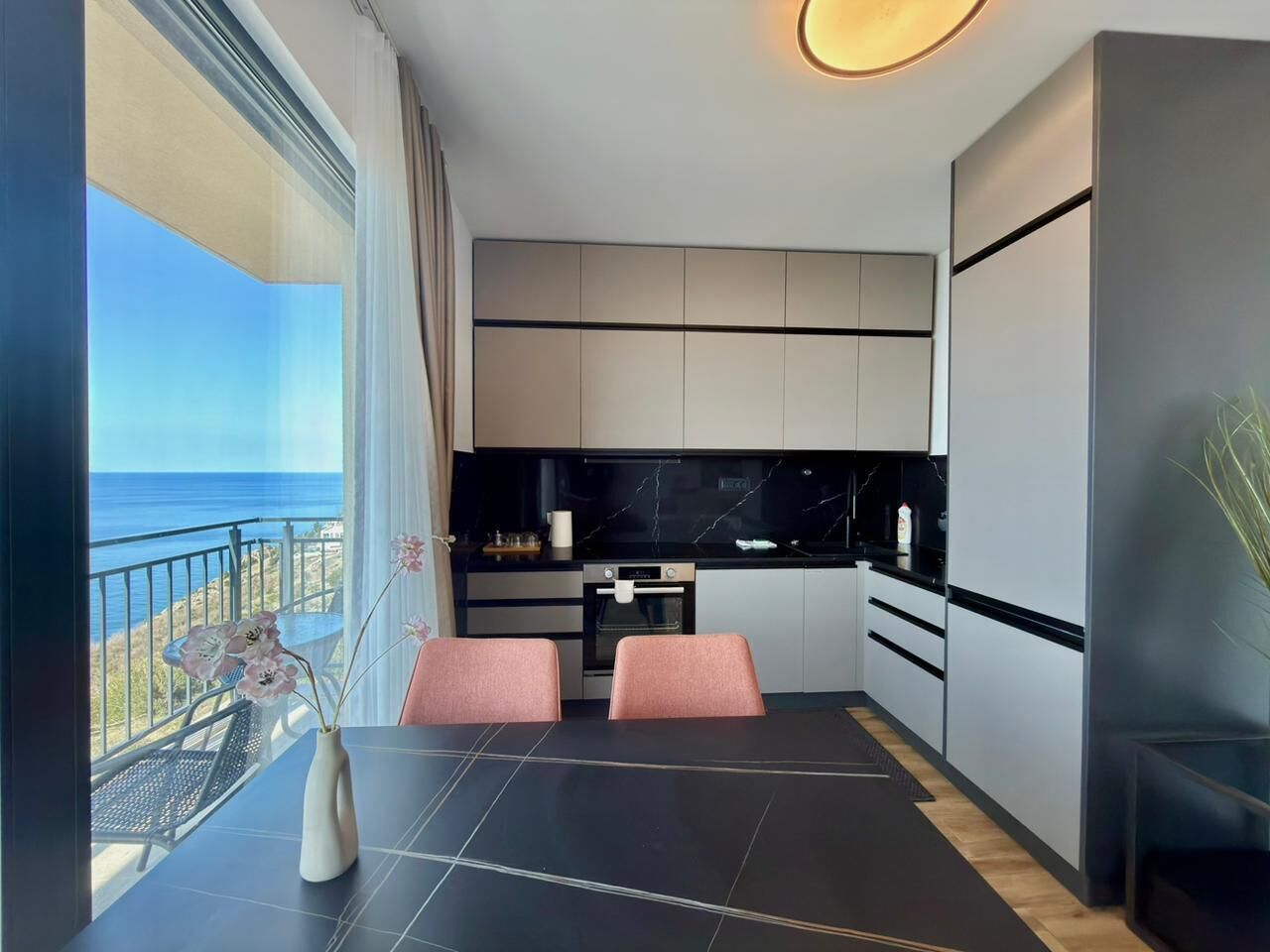 A modern kitchen with a view of the sea, a dining area with two pink chairs, and a large window.