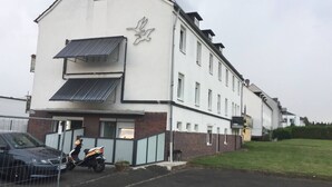 A two-story building with a white facade and a logo on the side. There is a parked car and a motorcycle in front. - NR 4 DG 2 rooms for companies and families (Nieder Vellmar)