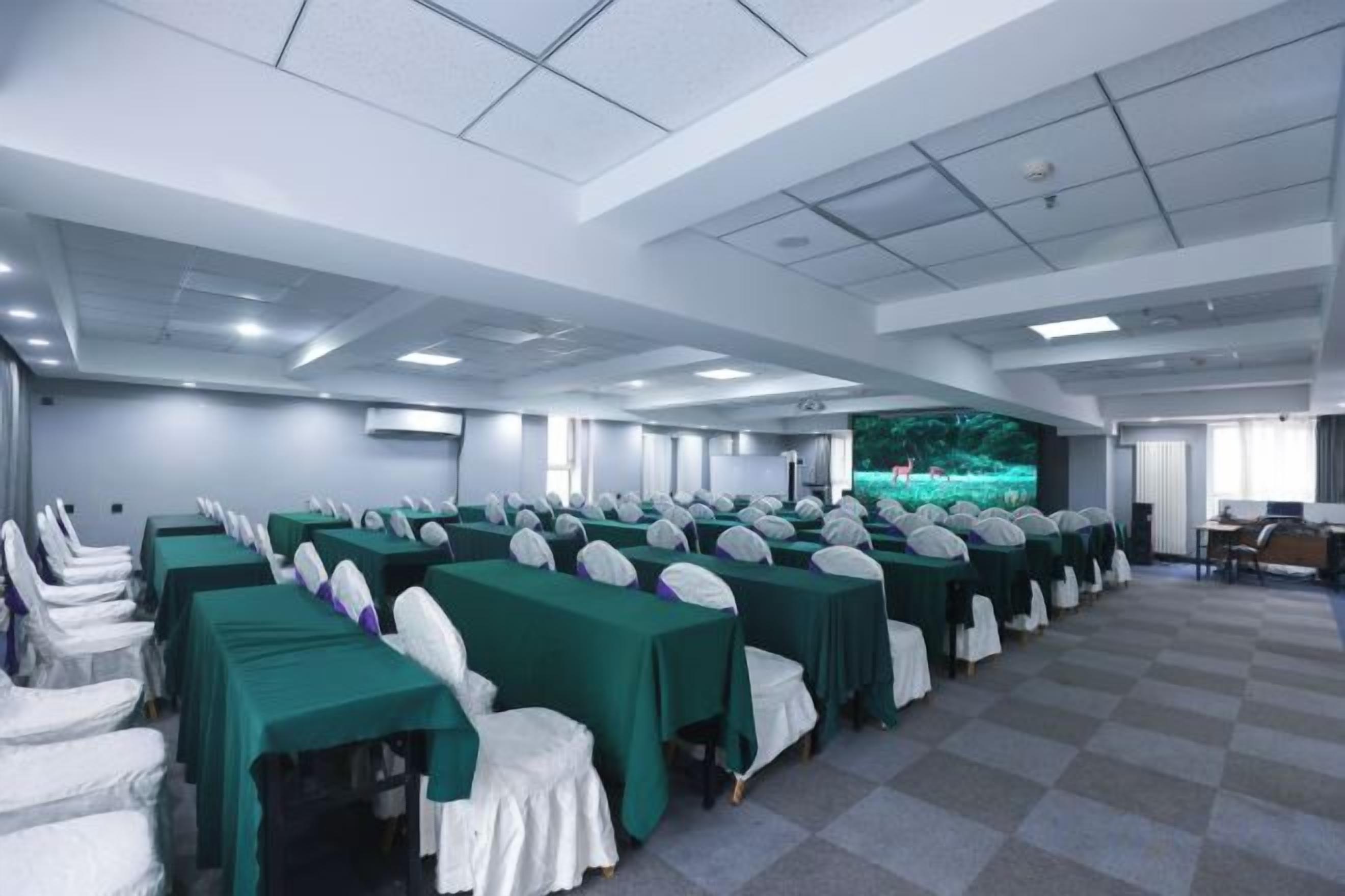 Une salle de conférence avec des rang ées de tables et de chaises, une scène dotée d’un écran de présentation et un sol à carreaux.