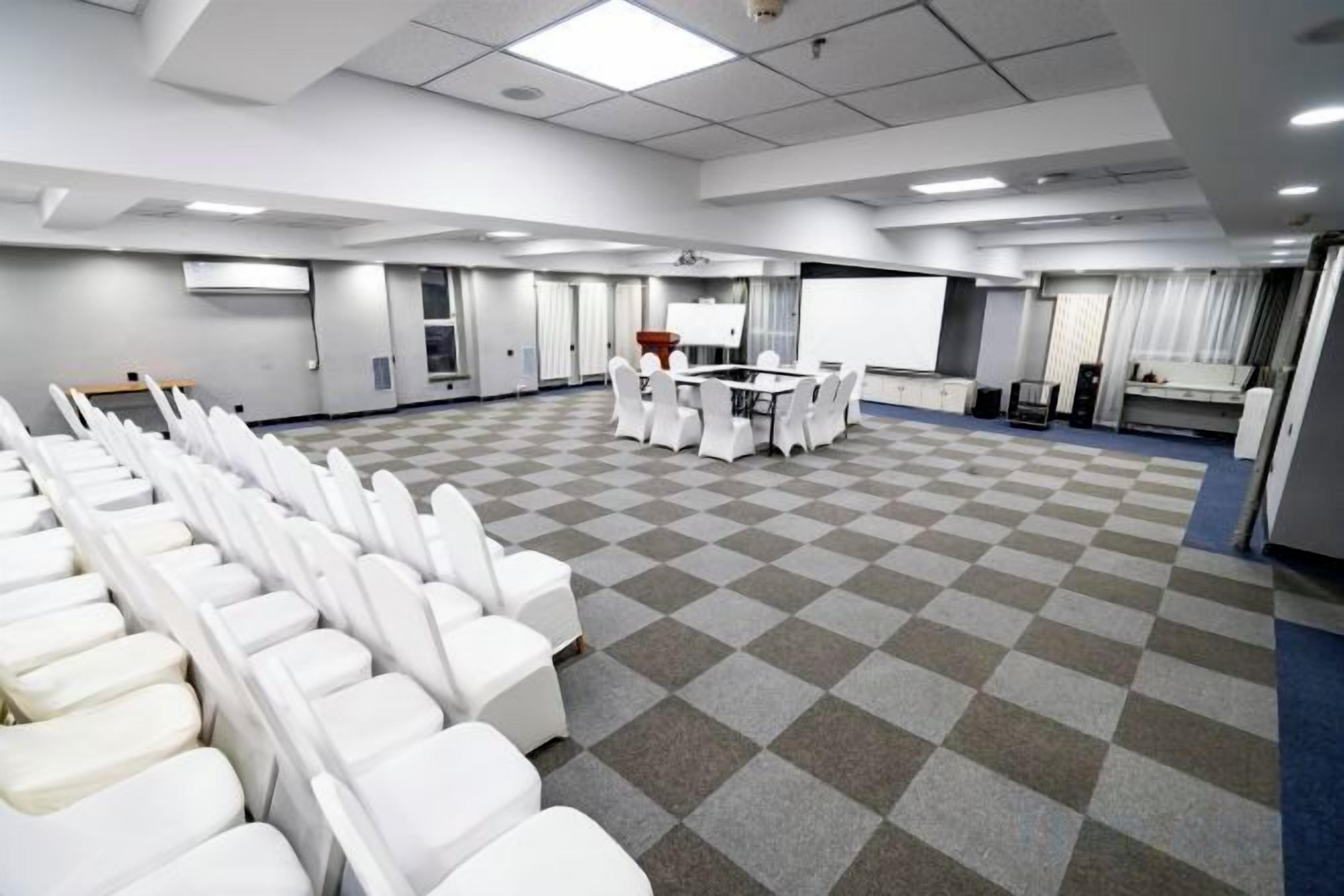 A conference room with rows of white chairs, a table with chairs, a projector screen, and a checkered floor.