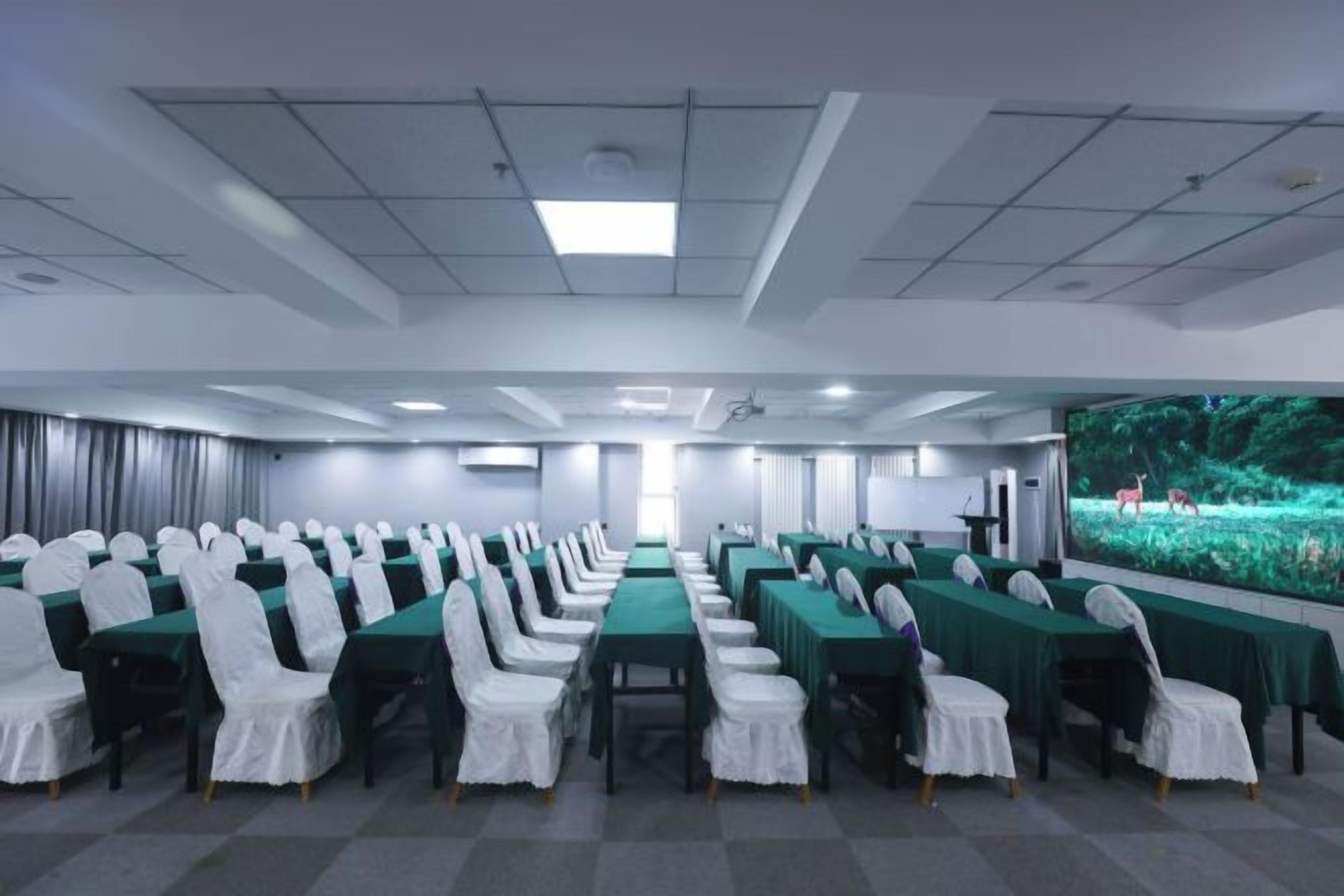 Un salón de conferencias con filas de mesas y sillas, una pantalla de proyector y un ventanal con vistas a zonas verdes.