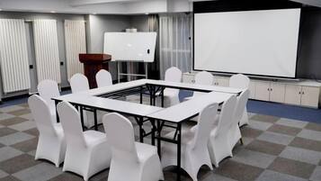 Un salón de conferencias con una gran mesa, sillas blancas, una pizarra y una pantalla de proyección.