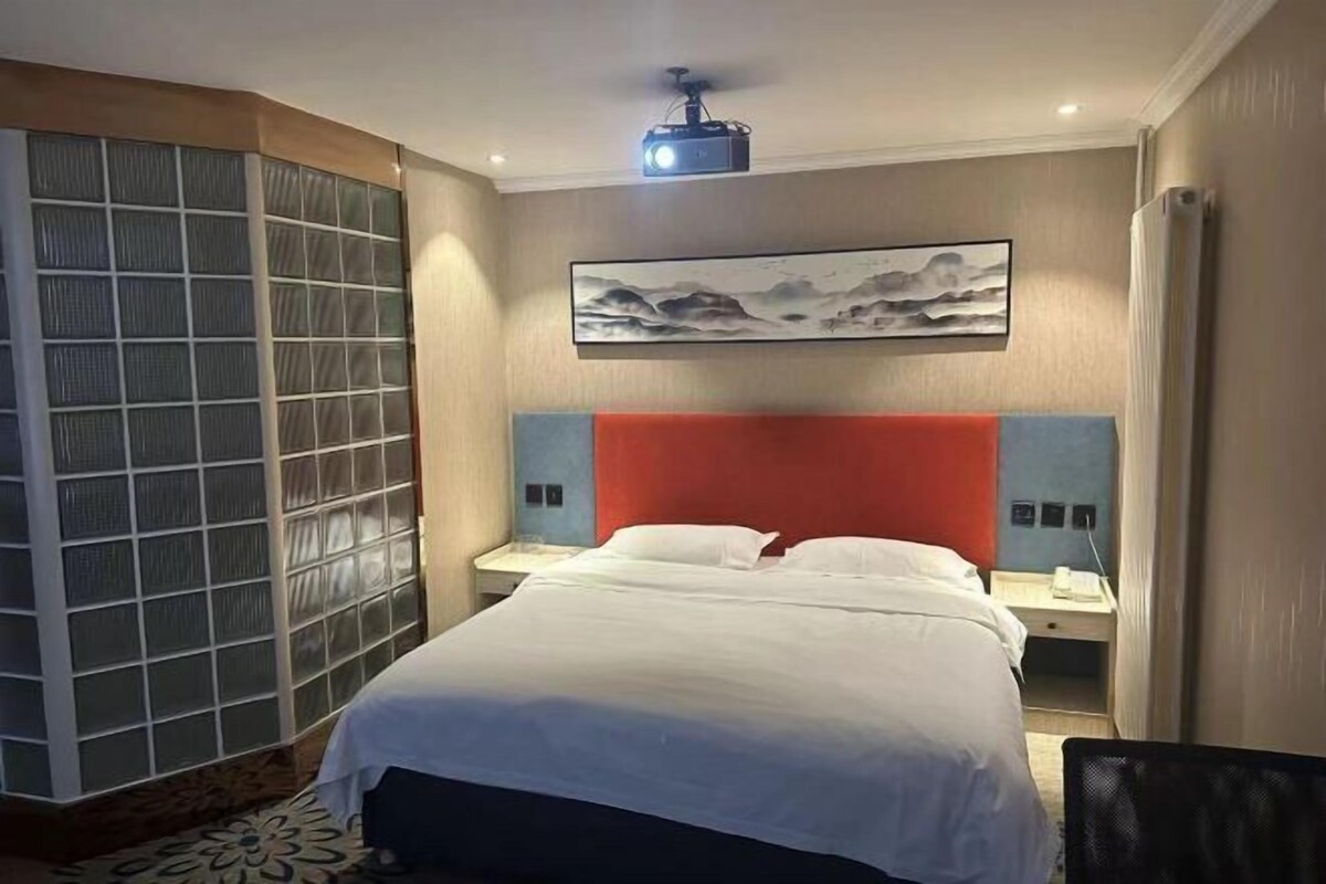 A hotel room with a bed, a nightstand, a projector, and a large window with a view of the mountains.
