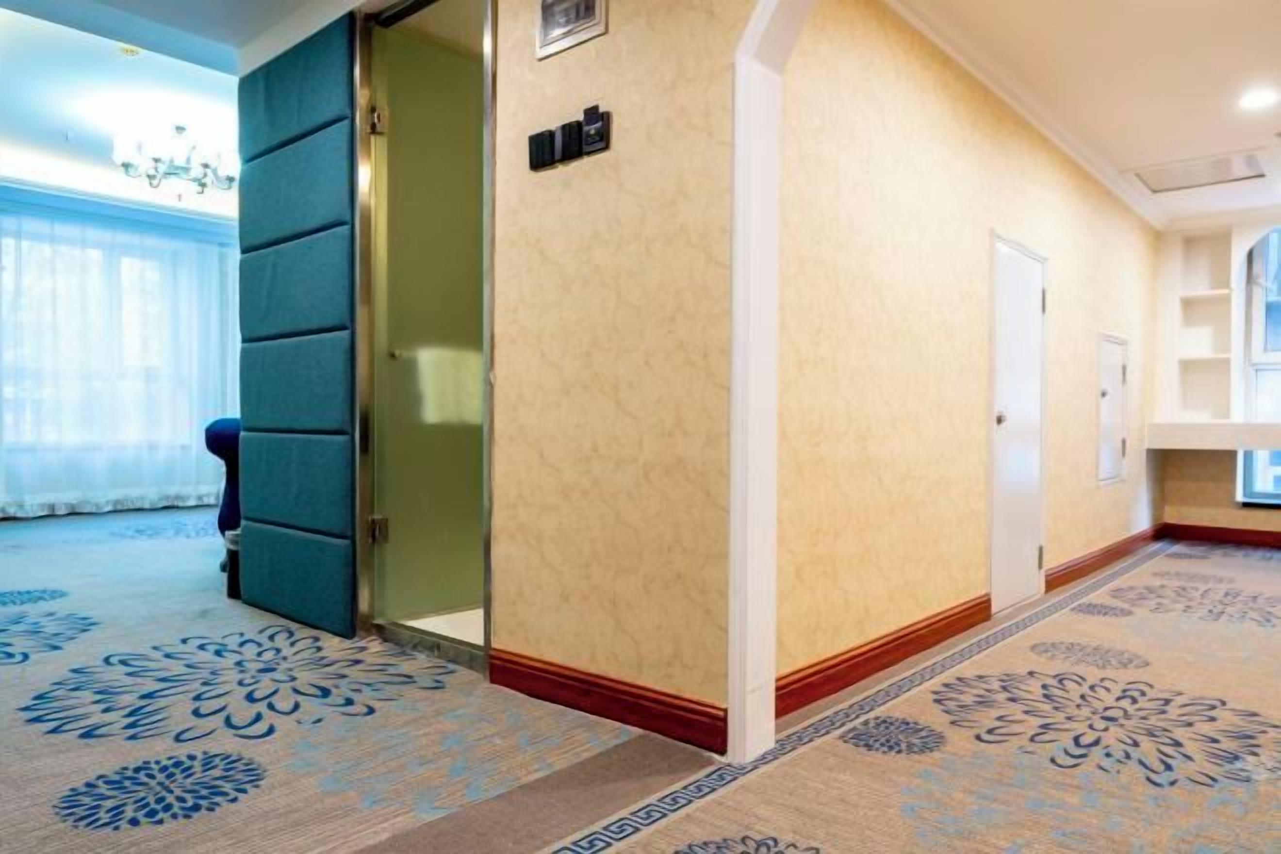 Un corridoio d'albergo con tappeto a motivi, una porta e una finestra con tende.