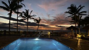 Une piscine entourée de palmiers, avec des chaises longues et des parasols, au moment du coucher du soleil.