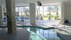 Una piscina interior con agua cristalina, rodeada de sillas de descanso y vistas a palmeras y un estacionamiento.
