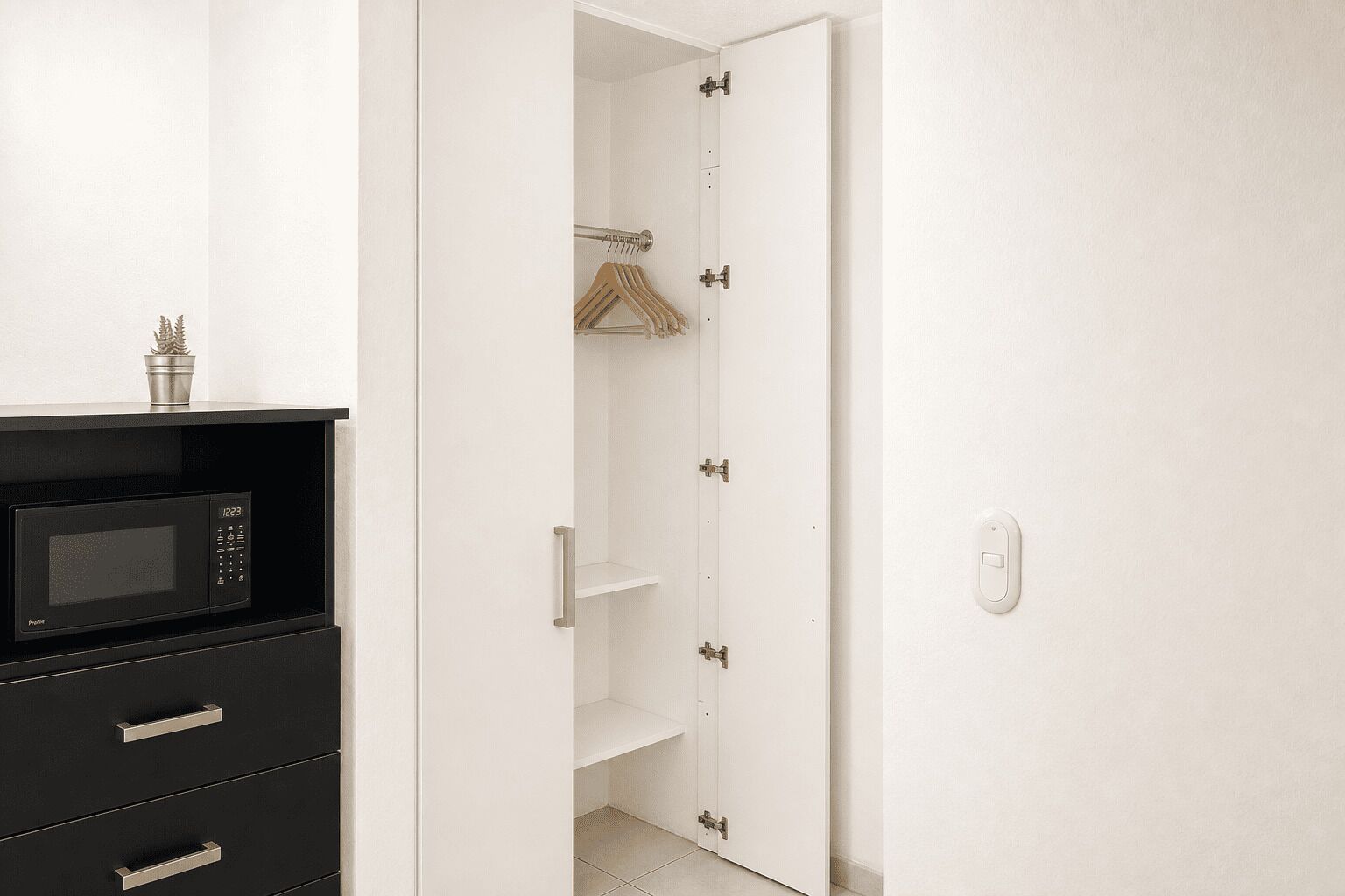 Una habitación con un microondas negro, una puerta blanca y un armario con ropa colgada.