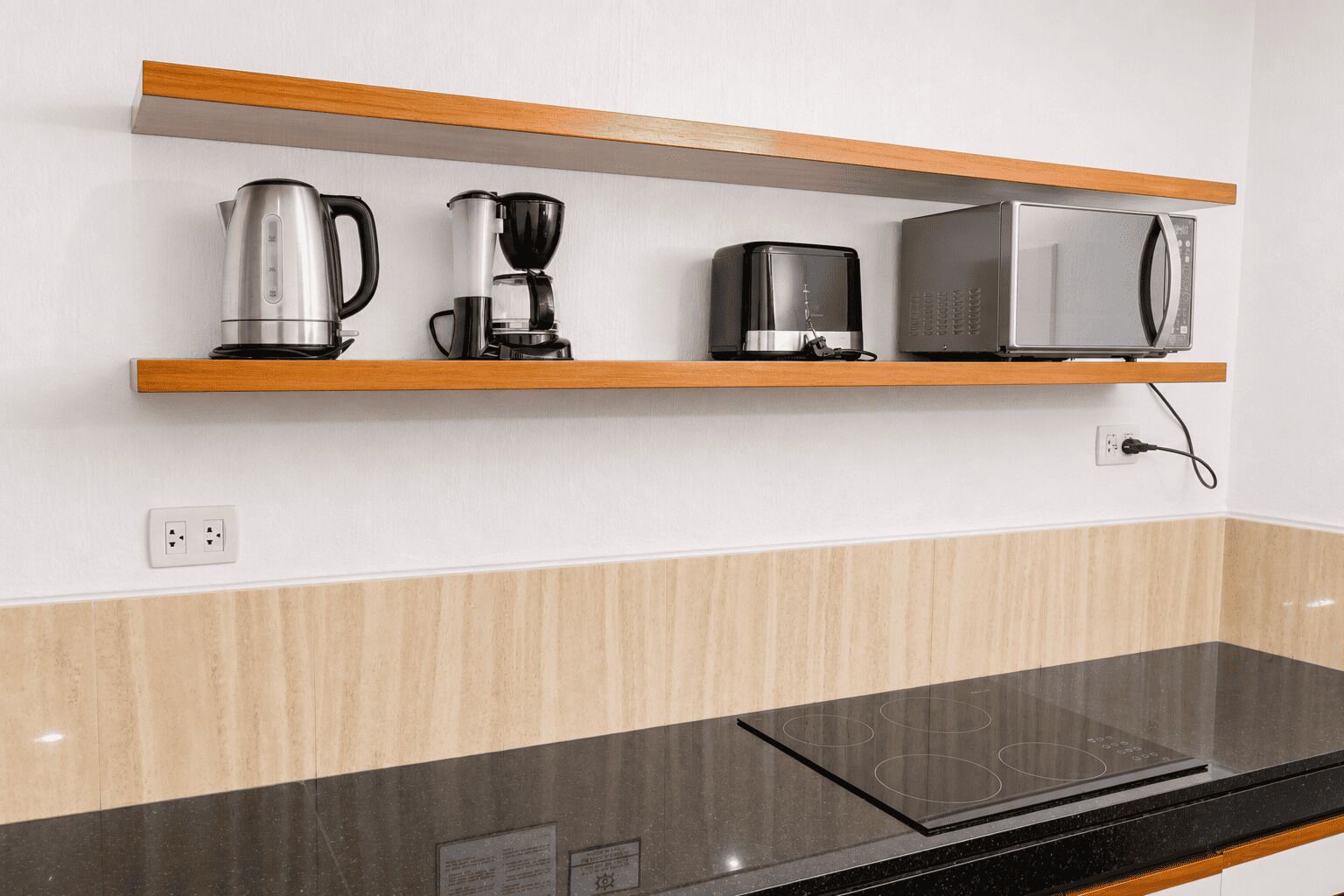 A kitchen with a black countertop, a stainless steel kettle, a coffee maker, and a microwave on a wooden shelf.