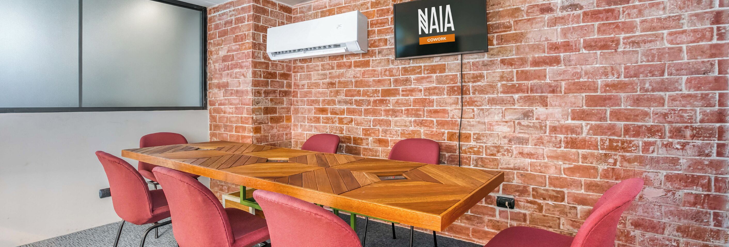 A conference room with a wooden table, red chairs, a brick wall, and a mounted TV screen.