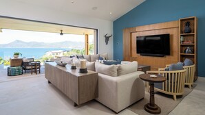 Una sala de estar moderna con un sofá grande, una mesa de centro y un televisor montado en una pared de madera. Se ve el mar a través de amplias ventanas.