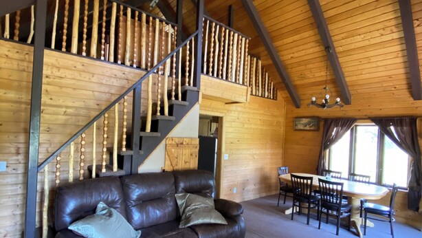 A spacious living area with a leather sofa, a dining table with chairs, a staircase with wooden balusters, and a high wooden ceiling.