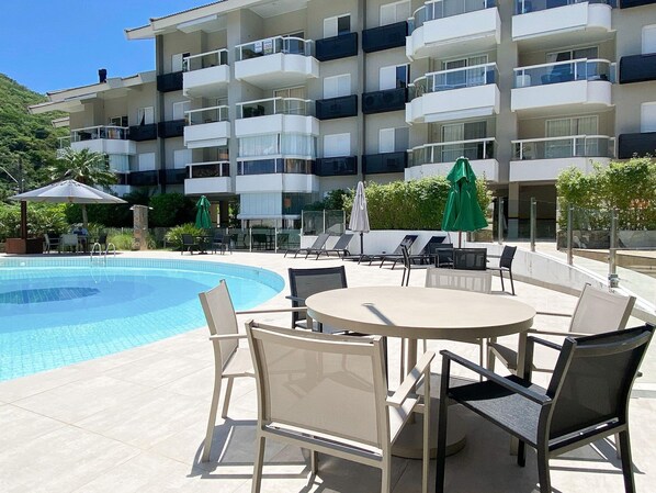 A poolside area with lounge chairs and umbrellas, a pool, and a multi-story building with balconies.