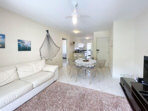 A living room with a white sofa, a dining table with chairs, a ceiling fan, and a television on a stand. - Residencial Porto da Brava (Florianopolis)