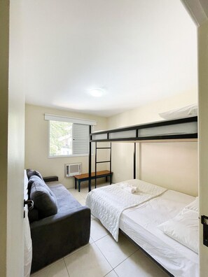 A dormitory room with bunk beds, a small table, a bench, and a window with blinds.
