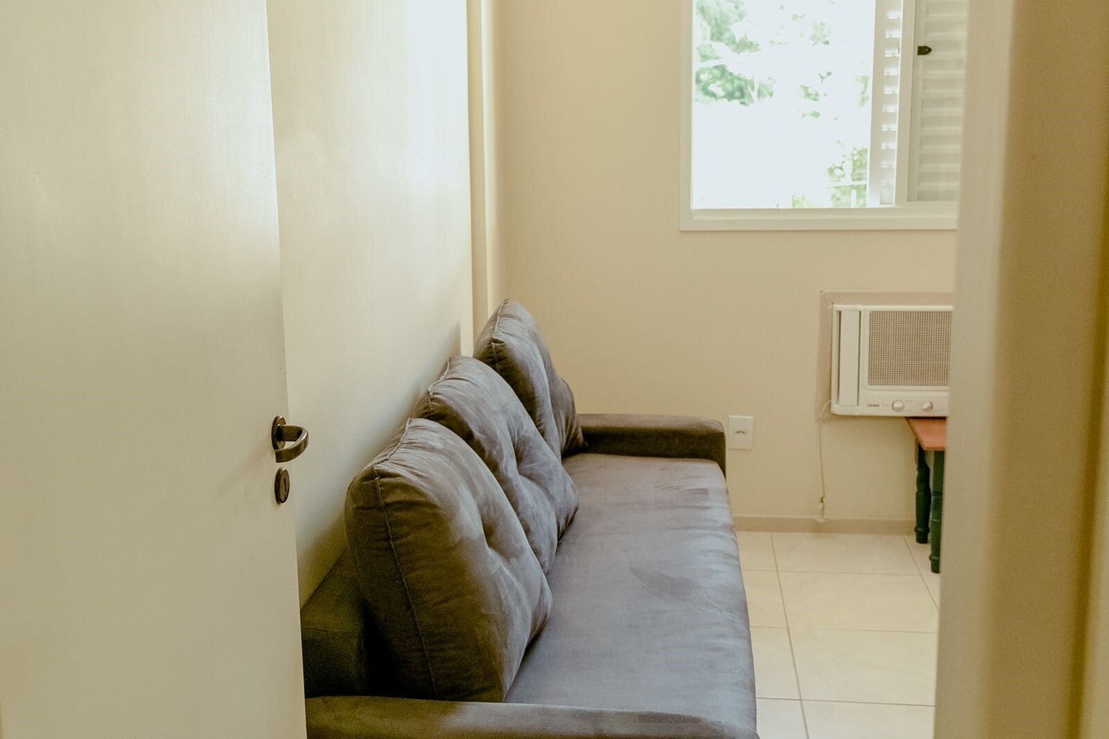 Una habitación con un sofá gris, una puerta blanca, una ventana con persianas y un aire acondicionado instalado en la pared.