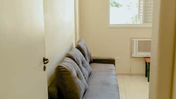 A room with a grey sofa, a white door, a window with blinds, and a wall-mounted air conditioner.