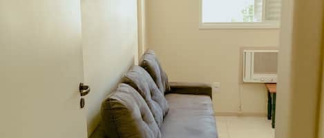 Una habitación con un sofá gris, una puerta blanca, una ventana con persianas y un aire acondicionado instalado en la pared.