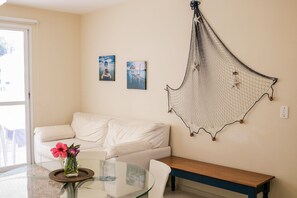 A modern living room with a glass-top coffee table, white sofa, and a wooden bench. There are two framed pictures on the wall and a decorative net hanging above. - Residencial Porto da Brava (Florianopolis)
