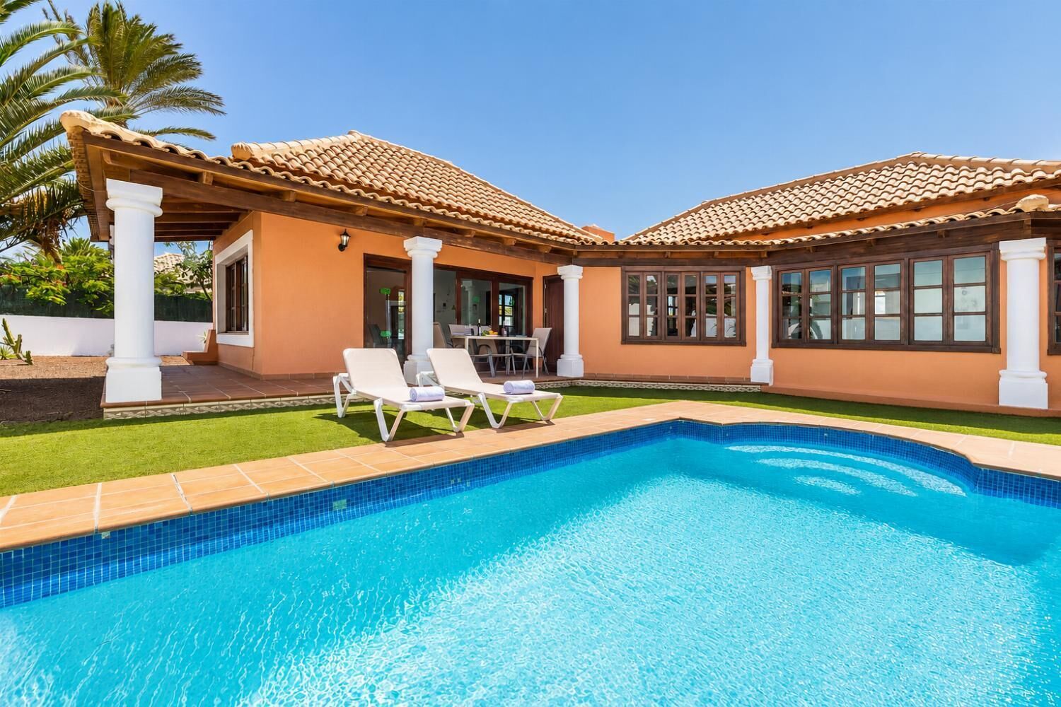 A swimming pool with lounge chairs, a covered patio, and a house with a tiled roof.