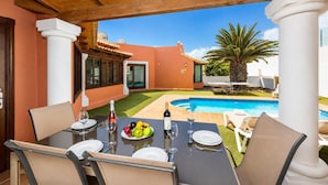 A dining area with a table set for two, a pool, and a view of a building with a tiled roof.