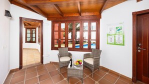 A room with two wicker chairs, a small table with a bottle and fruit, a wooden ceiling, a window with a view of a pool, and a door.